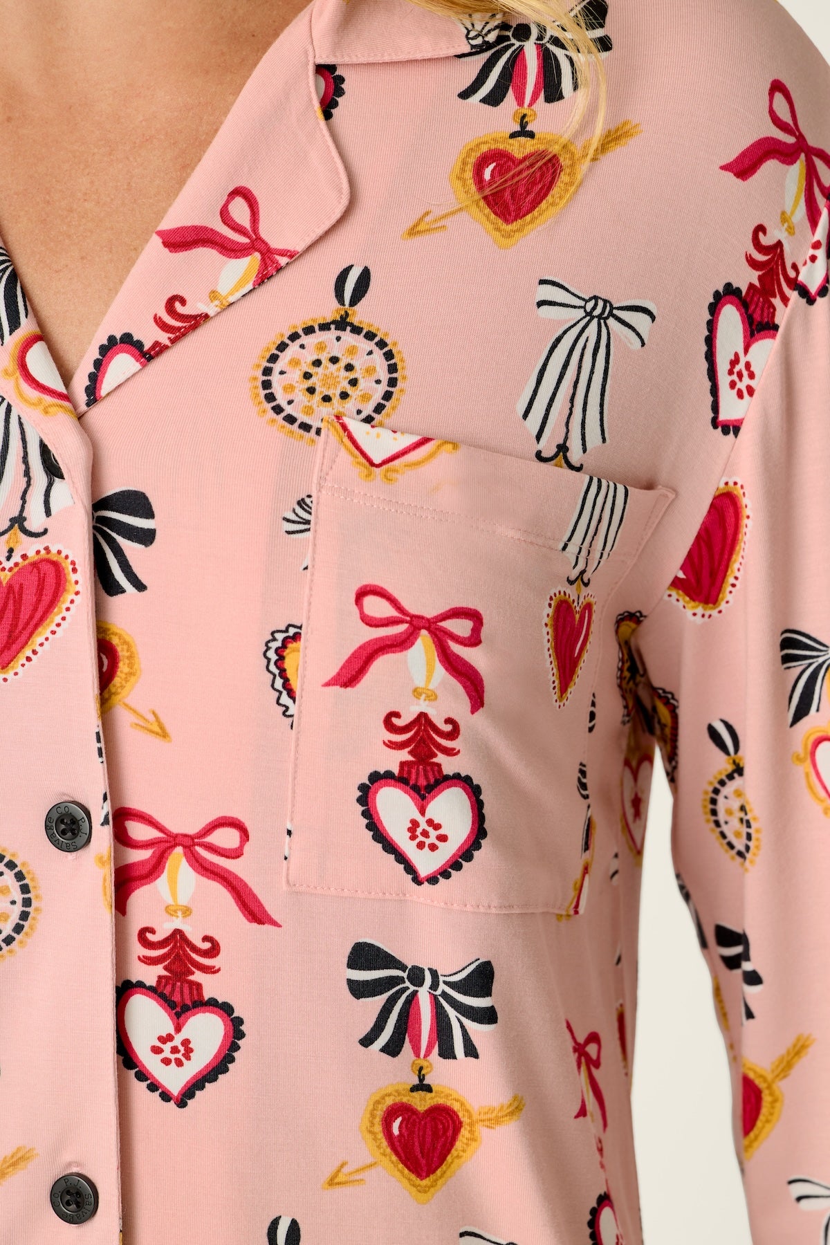 A close-up of a light pink pajama shirt with a repeating pattern of red, black, and yellow hearts, bows, and decorative ornaments, worn by a model.