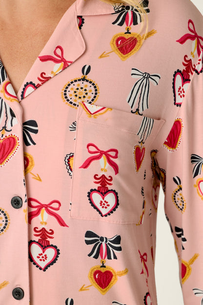 A close-up of a light pink pajama shirt with a repeating pattern of red, black, and yellow hearts, bows, and decorative ornaments, worn by a model.